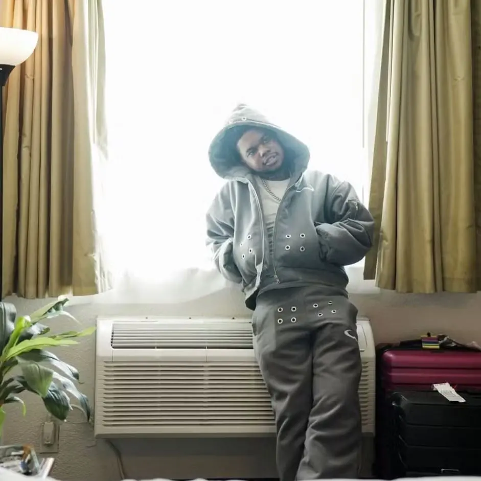 Pessoa vestida com um conjunto de moletom cinza com capuz, encostada em uma unidade de ar‑condicionado abaixo de uma janela com muita luz entrando. As cortinas são bege e há duas malas empilhadas ao lado da unidade, uma rosa e outra preta. À esquerda, há uma planta verde em um vaso. A pessoa está com as mãos nos bolsos e as pernas cruzadas.