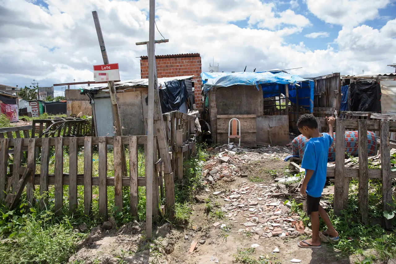 Imagem mostra Ocupação Terra Prometida, comunidade carente com casas improvisadas em Fortaleza, com foco em crianças brincando na rua e céu parcialmente nublado, representando dificuldades de moradia e condições precárias.