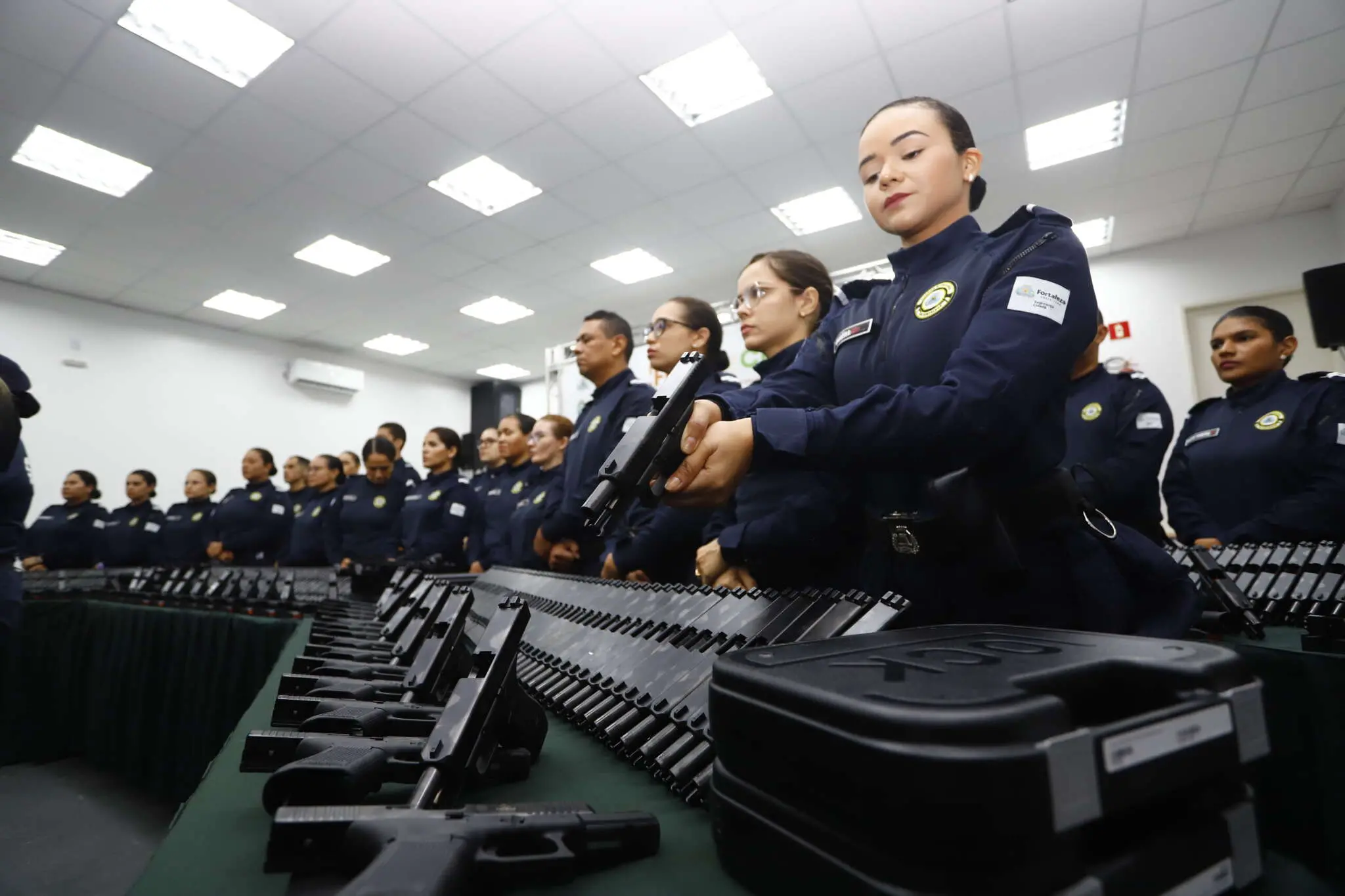 Grupo de agentes uniformizados durante cerimônia de entrega de armamentos, segurando pistolas e posicionados ao lado de várias armas organizadas sobre uma mesa em sala iluminada. A foto é de uma cerimônia da guarda municipal de fortaleza recebendo novas armas.