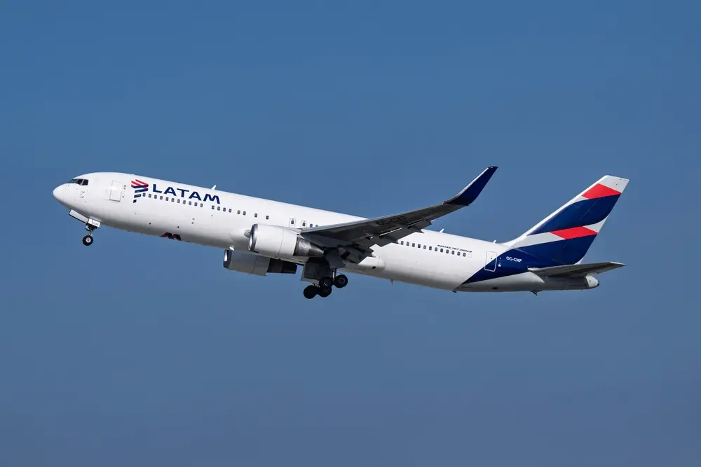 Avião branco da companhia LATAM em voo, visto de lado, contra um céu azul. A aeronave está inclinado levemente para cima, com o trem de pouso recolhido e as asas estendidas. A cauda exibe as cores azul, vermelho e branco da marca.