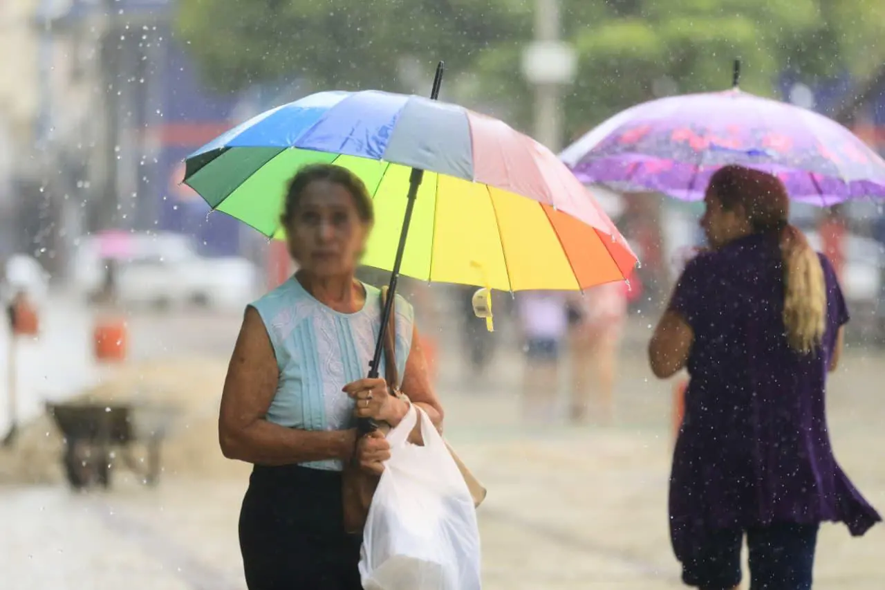 Mulher caminhando sob chuva forte, segurando um guarda-chuva arco-íris vibrante em uma rua urbana molhada, com outras pessoas ao fundo.