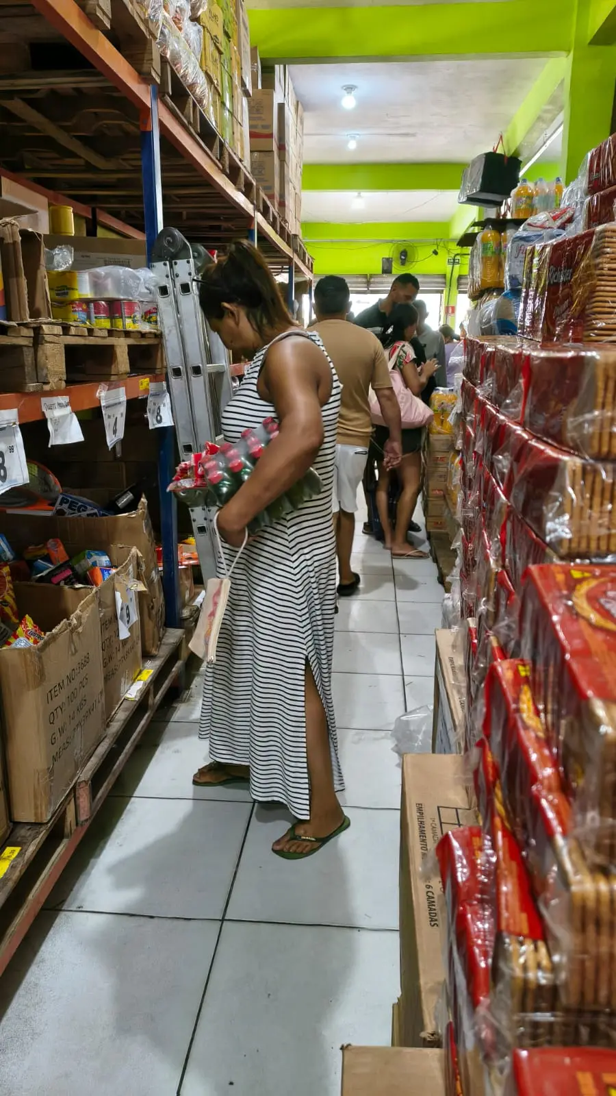 Cena de compras em um supermercado ou armazém, com uma mulher em primeiro plano examinando produtos nas prateleiras e outras pessoas ao fundo.