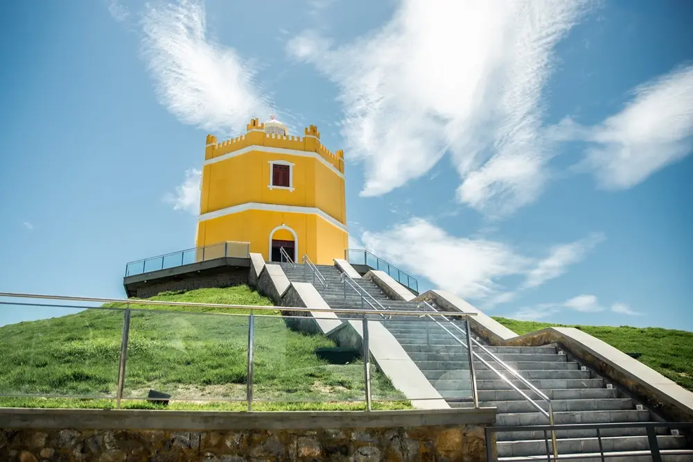 Na imagem, o Farol do Mucuripe, localizado em Fortaleza, Ceará. O edifício histórico possui um formato octogonal e é pintado em um amarelo vibrante, com detalhes em branco nas molduras e janelas. Ele está situado no topo de uma pequena colina gramada, sob um céu azul intenso com nuvens brancas finas e alongadas que parecem irradiar de trás da estrutura. No primeiro plano, há uma escadaria de pedra cinza com corrimãos metálicos que leva até a entrada do farol. A base do terreno é cercada por um guarda-corpo de vidro moderno. A iluminação é de luz do dia natural, realçando o contraste entre o amarelo do prédio, o verde da grama e o azul do céu.
