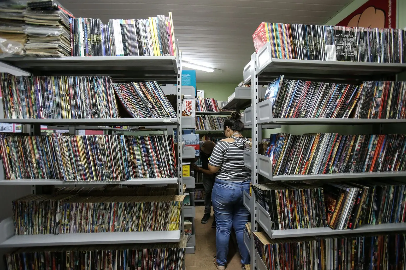Estantes repletas de gibis e quadrinhos na Biblioteca Dolor Barreira, com duas pessoas navegando pela coleção.