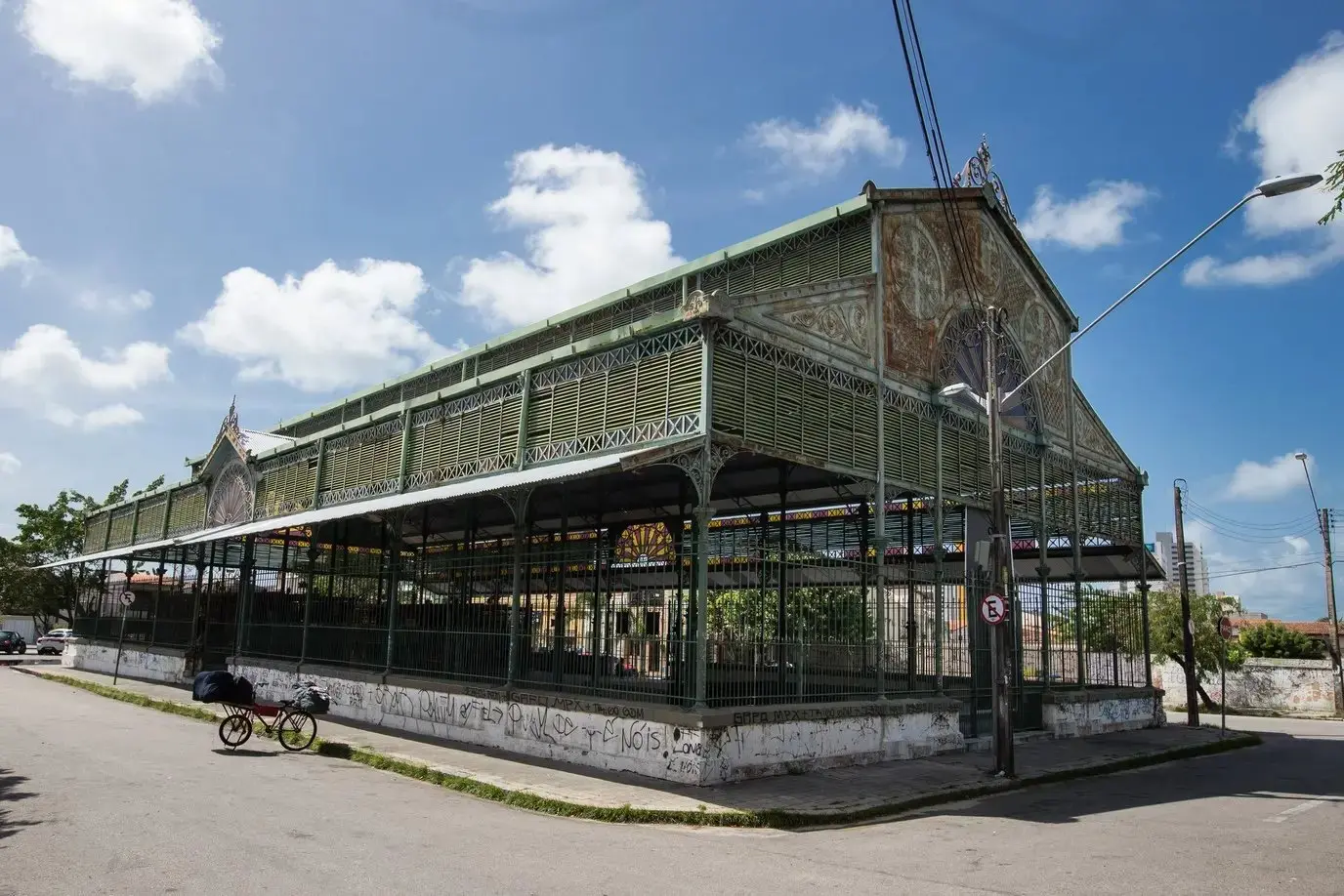 Vista panorâmica de um mercado público antigo, o Mercado dos Pinhões, com estrutura de ferro verde e detalhes ornamentados, sob um céu azul claro.