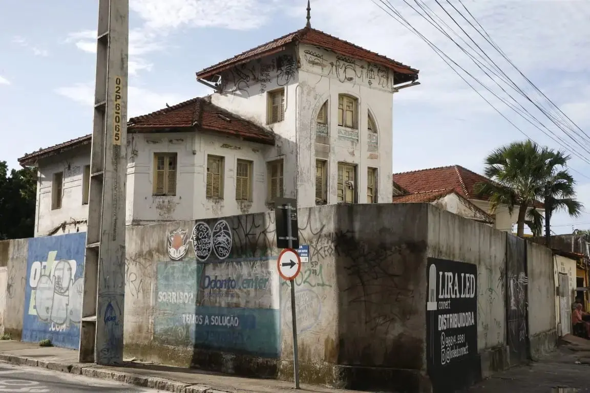Antiga casa de arquitetura eclética com telhados de cerâmica e fachada desgastada, cercada por um muro pichado com anúncios urbanos, sob um céu claro e fios elétricos.