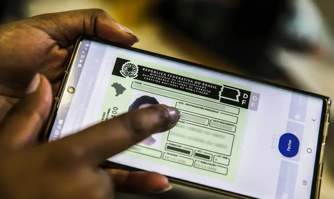 Uma pessoa segura um smartphone que exibe a Carteira Nacional de Habilitação (CNH) digital em tons de verde e branco. O dedo indicador toca a tela sobre os dados do documento, destacando a modernidade do acesso à identificação em um ambiente de luz suave.