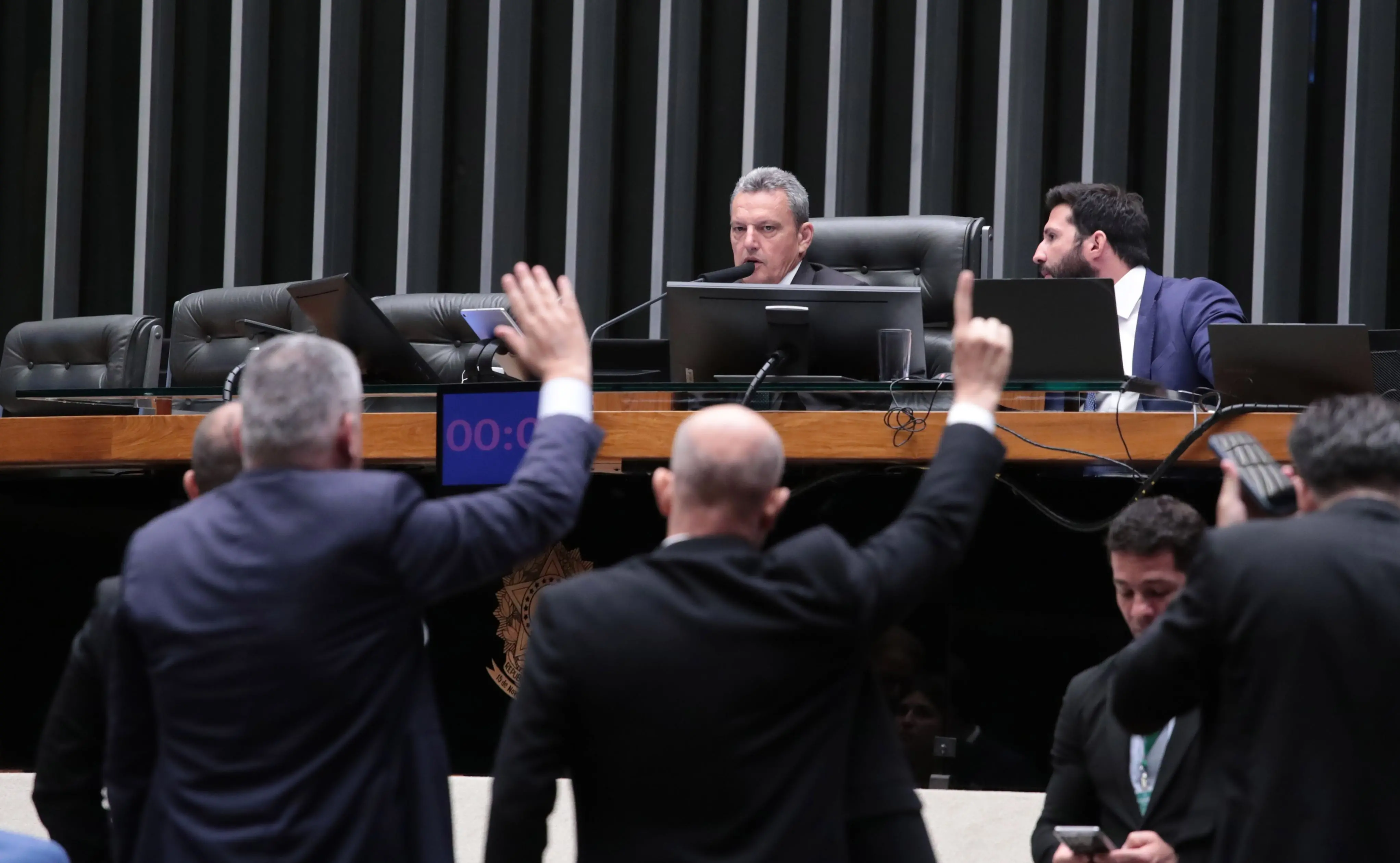 Parlamentares de terno levantam a mão para votar no plenário da Câmara, enquanto o presidente da sessão, sentado à mesa diretora com microfone e monitores, conduz os trabalhos.