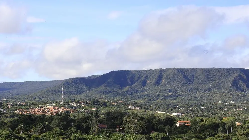 Fotografia panorâmica de uma encosta de serra densamente arborizada sob um céu azul com nuvens brancas. No sopé da elevação, destaca-se uma área urbana com casas de telhados cerâmicos e uma torre de comunicação alta e fina. A vegetação é verde vibrante, típica de regiões tropicais, cobrindo quase toda a extensão do relevo acidentado.