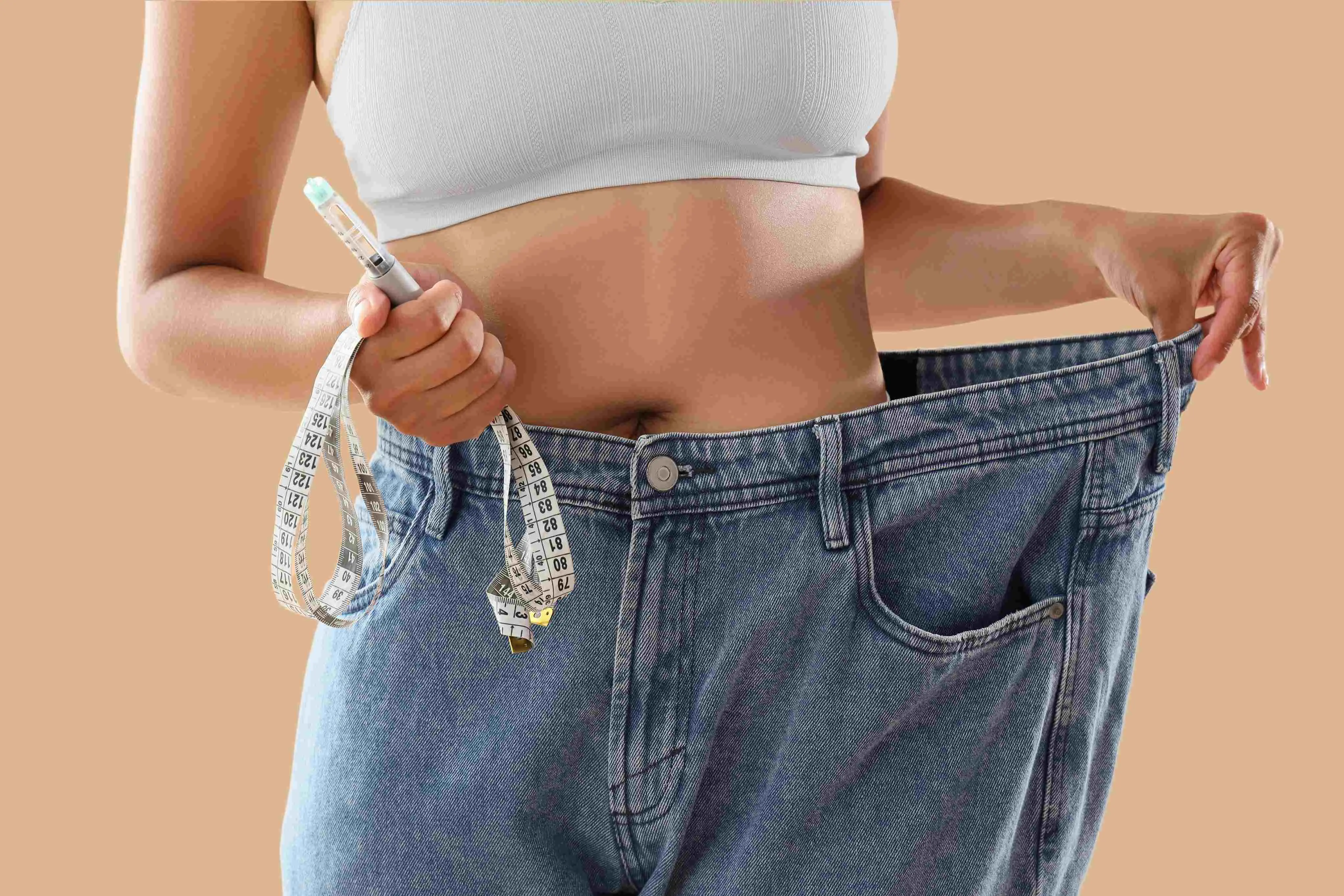 Mulher vestindo calças jeans visivelmente largas e segurando uma fita métrica e uma caneta injetável de medicação, simbolizando o processo de emagrecimento.