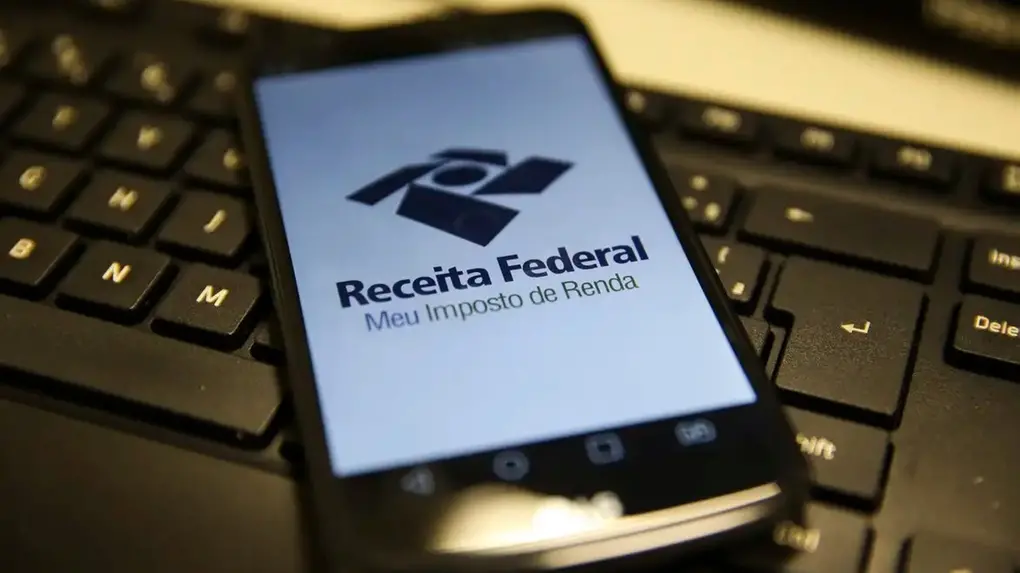 Smartphone exibindo a tela do aplicativo “Receita Federal – Meu Imposto de Renda” apoiado sobre um teclado de computador.