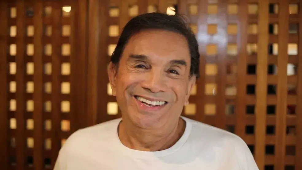 Homem sorridente com cabelo escuro e roupa branca, fundo de madeira, transmitindo felicidade e bem-estar.