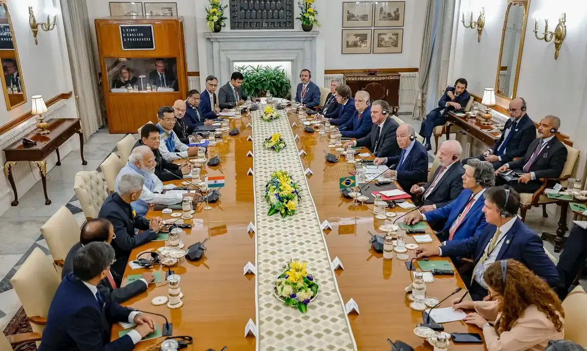 Reunião internacional de líderes e representantes de diferentes países tomando decisões importantes em uma sala de convenções com decoração elegante, flores na mesa e painel de vídeo ao fundo.
