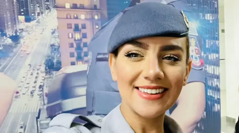 Agente de segurança feminina sorrindo com uniforme e capacete, em ambiente urbano com prédio ao fundo.