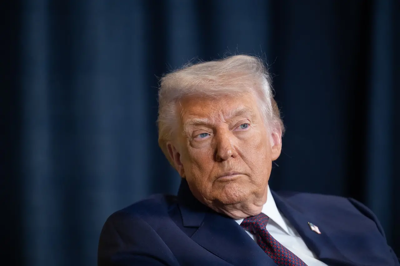 Donald Trump, homem de terno azul e gravata vermelha, com expressão séria, sentado diante de um fundo escuro desfocado.