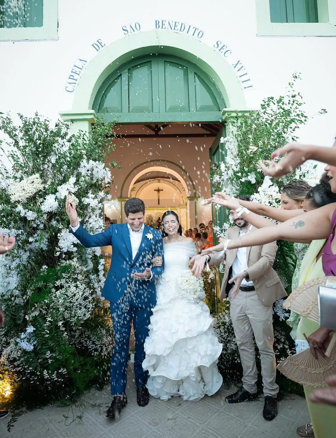 Na imagem, uma foto colorida mostra um casal de noivos saindo de uma igreja histórica sob uma chuva de arroz. No centro, o noivo veste um terno azul vibrante e sorri, enquanto a noiva, ao seu lado, usa um vestido branco de ombros de fora com uma saia volumosa em camadas de babados. Ela segura um buquê de flores brancas e sorri com os olhos fechados. Eles estão saindo pela porta verde de arco da 
