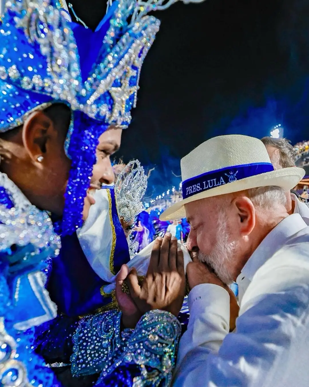 Lula beija bandeira de escola de samba durante carnaval do Rio. Ele está com um chapéu e uma faixa azul e veste uma roupa branca.
