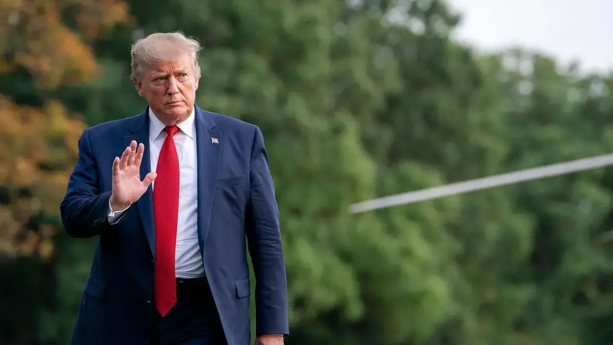 Trump caminha e acena em um gramado. Ele está de paletó e uma gravata vermelha. Ele parece olhar para o horizonte enquanto se desloca.