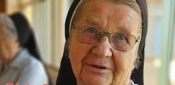 Close de uma freira idosa com hábito preto e véu, usando óculos e olhando levemente para a câmera, com expressão serena; outra religiosa aparece desfocada ao fundo.