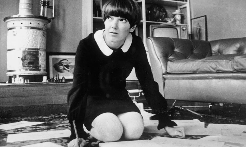 Na imagem, fotografia em preto e branco da estilista Mary Quant nos anos 60. Ela está ajoelhada no chão sobre papéis de desenho, segurando um lápis. Mary usa seu icônico corte de cabelo 