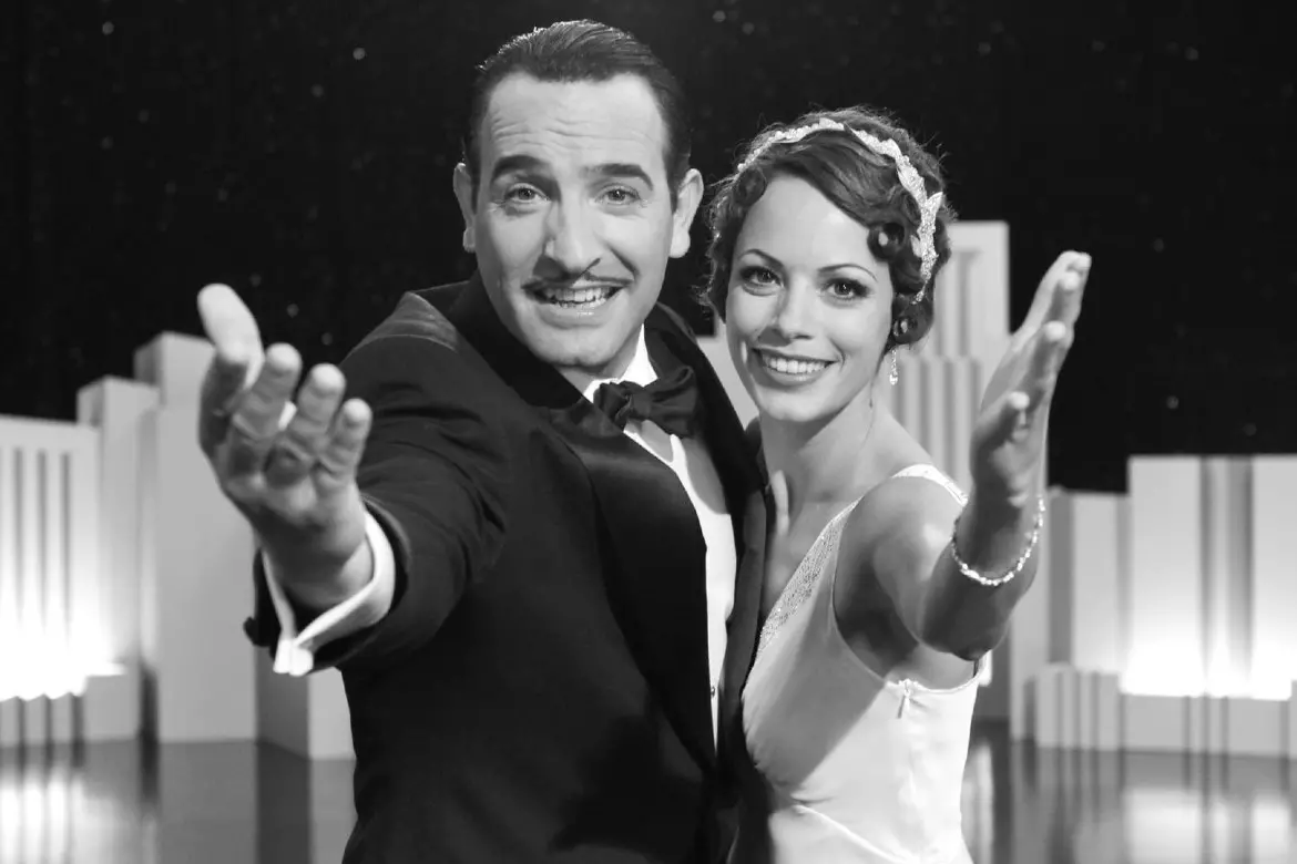 Casal em elegância em um evento, vestido formalmente, sorrindo e com postura confiante, no cenário de um palco ou um set de filmagem, em preto e branco, promovendo felicidade e sofisticação.