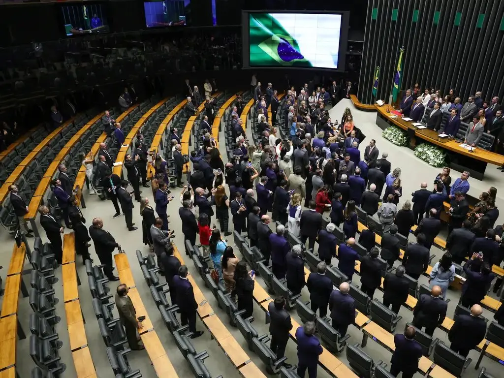 Imagem aérea do interior do Congresso Nacional, com parlamentares em pé, trajando terno, com imagem de bandeira do Brasil reproduzida em projetor.