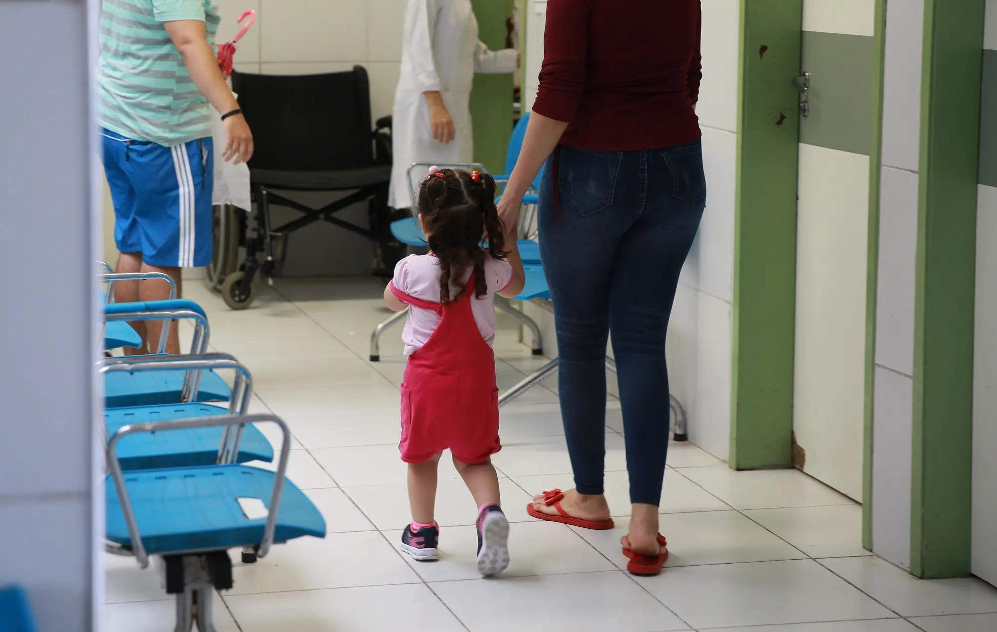 Mãe e filha caminhando em uma área de espera médica, com outros pacientes e profissionais de saúde no entorno.