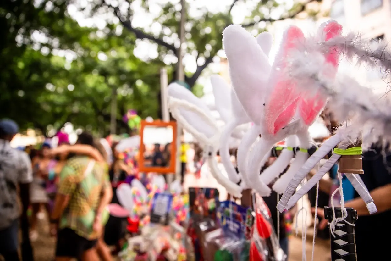 Acessórios de fantasia, como tiaras de orelhas de coelho felpudas, à venda em um evento ao ar livre animado, com a multidão e itens festivos em segundo plano.