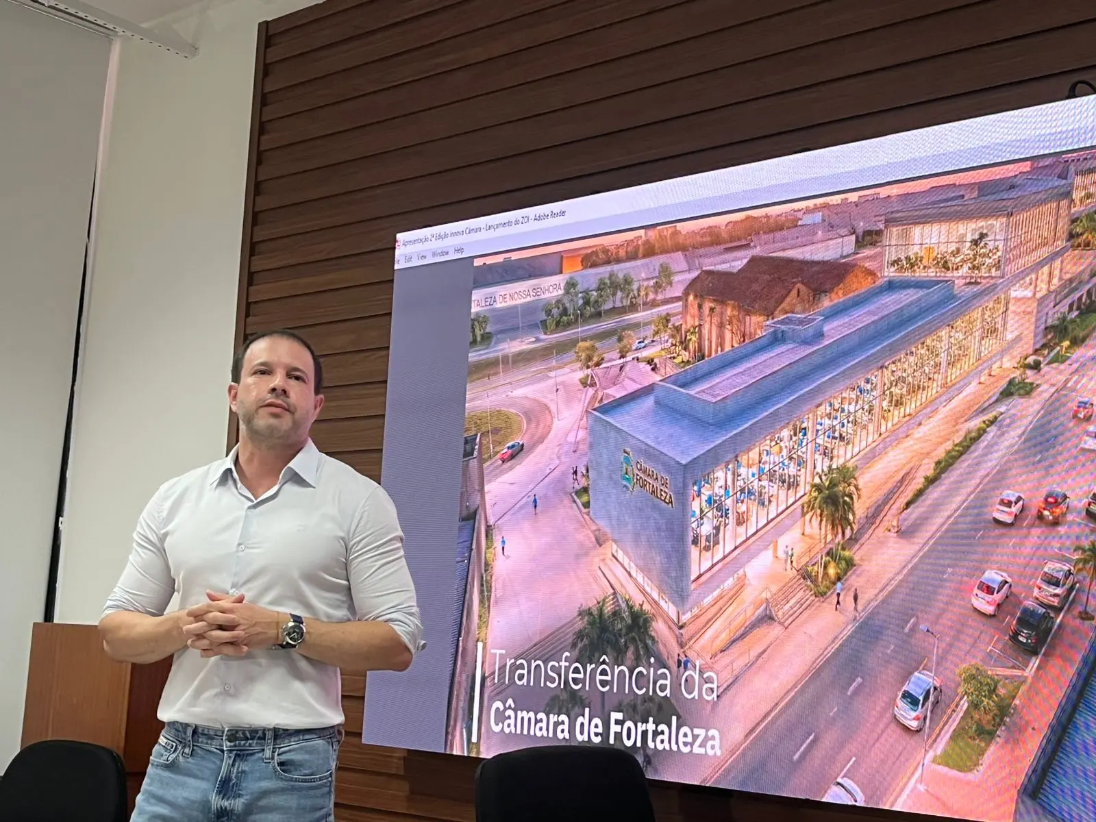 Léo Couto, presidente da Câmara Municipal de Fortaleza, de camisa branca e jeans apresenta projeto ao lado de tela que exibe imagem da nova sede da Câmara de Fortaleza e o texto ‘Transferência da Câmara de Fortaleza’, em sala com parede de madeira.