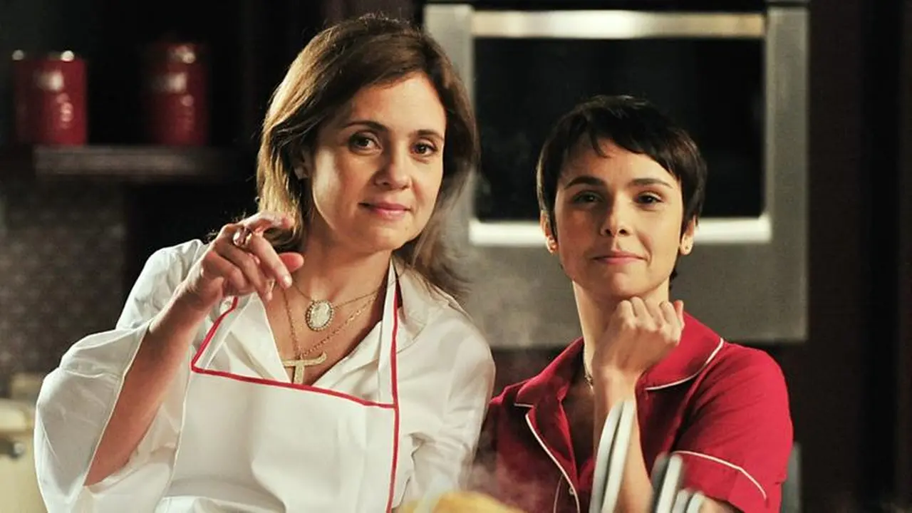 As atrizes Adriana Esteves e Débora Falabella posam caracterizadas como Carminha e Nina em uma cozinha, com Carminha apontando para a câmera enquanto veste um avental branco. O cenário de fundo apresenta utensílios domésticos e um forno, capturando a rivalidade icônica das personagens em uma atmosfera cotidiana e expressiva.