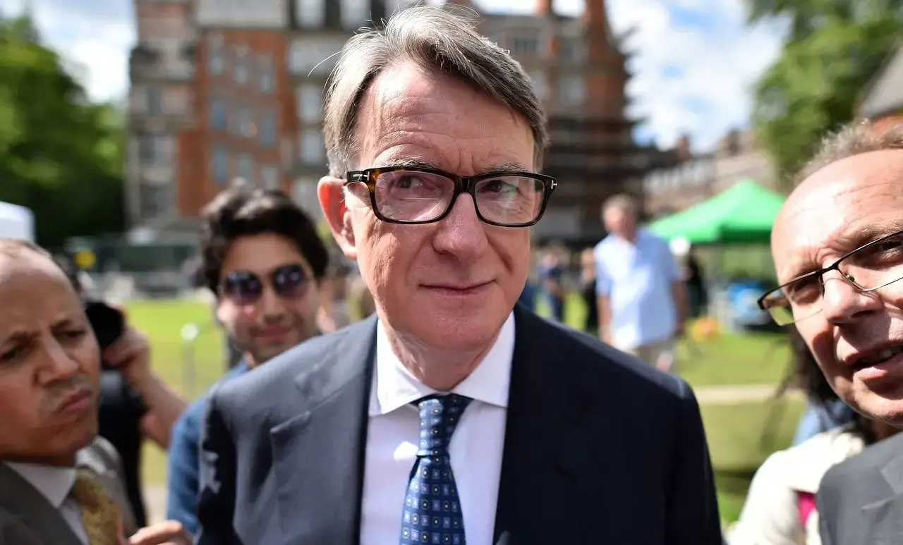 Ex-embaixador britânico Peter Mandelson, que usa óculos de armação escura, terno azul e gravata padronizada, aparece em um close centralizado enquanto é cercado por outras pessoas e fotógrafos. Ao fundo, um gramado ensolarado e edifícios de tijolos vermelhos compõem o cenário externo sob uma iluminação natural. Imagem usada em matéria sobre o caso Epstein.