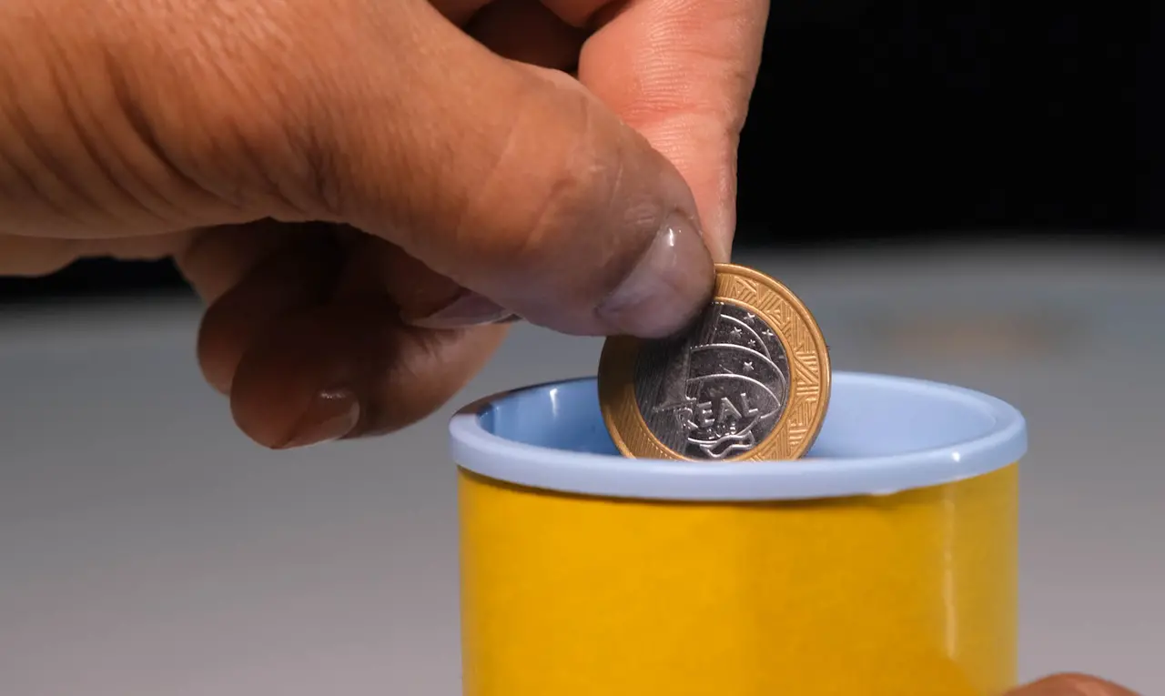 Mão segurando uma moeda de um real e colocando-a dentro de um pequeno recipiente amarelo com borda azul, sobre uma superfície clara e com fundo escuro desfocado.