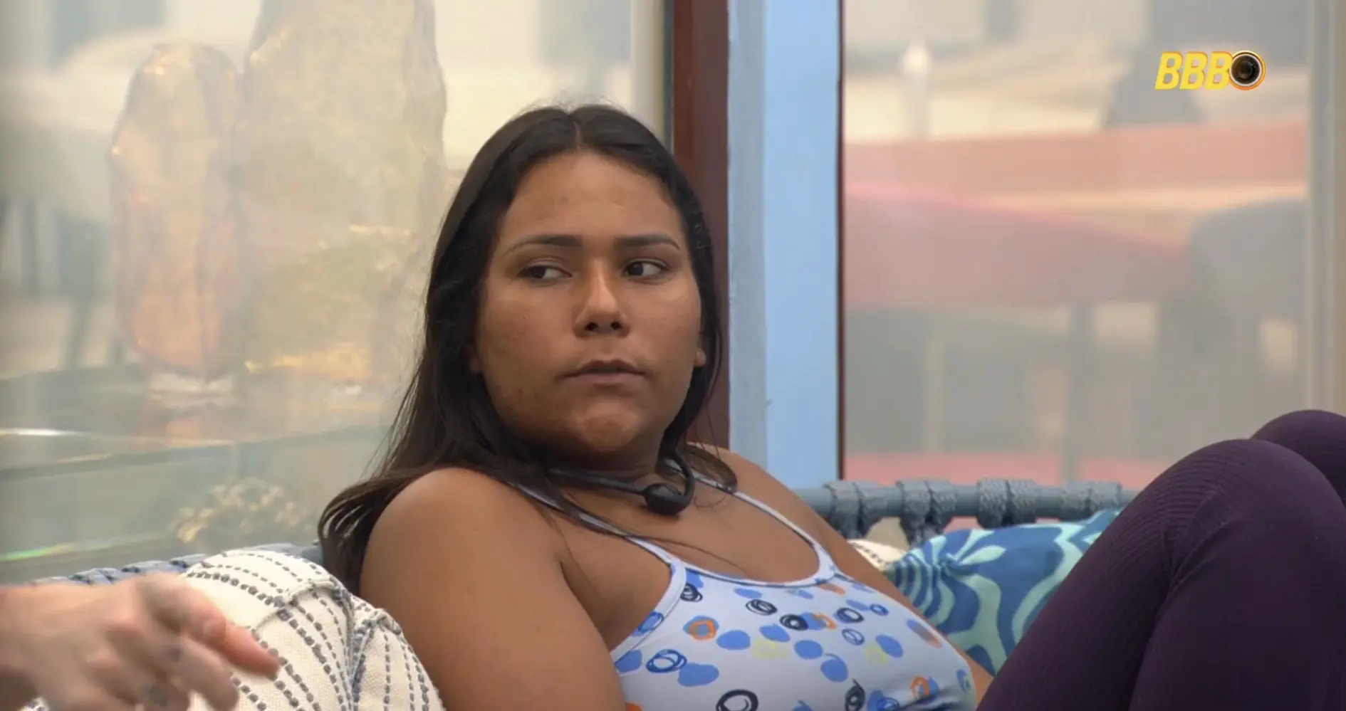 Mulher de cabelo escuro e expressão séria sentada em sofá, dentro de um ambiente bem iluminado, com fundo de vidro e decoração moderna, em cena de reality show.