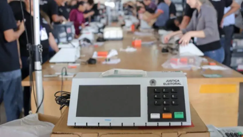 Urna eletrônica da Justiça Eleitoral sobre mesa de papelão em primeiro plano, com pessoas ao fundo realizando procedimentos de preparação dos equipamentos em um ambiente interno.