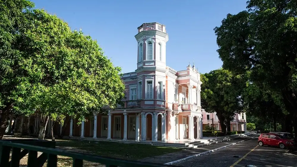 Imagem do edifício histórico da Universidade Federal do Ceará (UFC) com arquitetura clássica, jardins bem cuidados, árvores e céu azul ao fundo, destacando a beleza e tradição da instituição.
