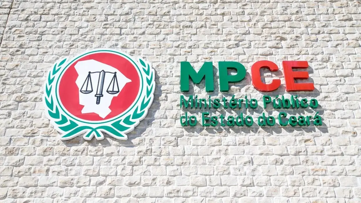 O logotipo do Ministério Público do Estado do Ceará está fixado em uma parede de pedras claras, exibindo um brasão circular com o mapa do estado e a balança da justiça. Ao lado, a sigla MPCE em relevo verde e vermelho acompanha o nome da instituição em letras verdes, criando uma composição institucional sóbria e oficial.