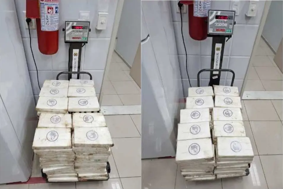 “Pilha de tabletes brancos embalados, contendo cocaína, com selos circulares impressos, colocados sobre uma balança em ambiente interno com piso claro e extintor de incêndio ao fundo.