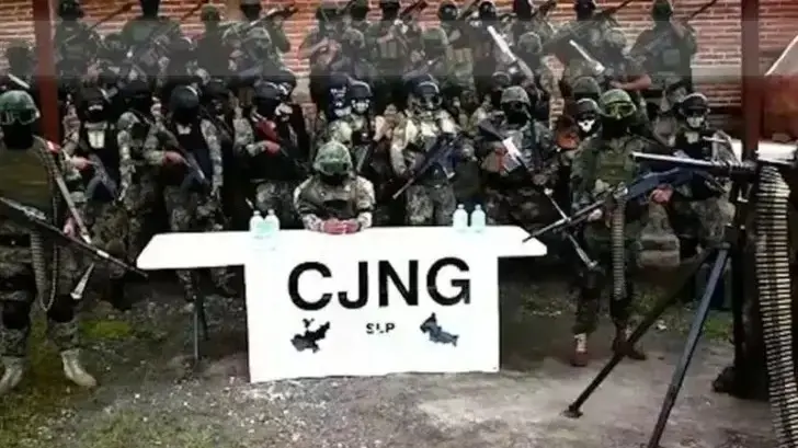 A foto mostra vários homens vestidos como militares e armados com armas de guerra, posando para foto com uma bandeira do CJNG.