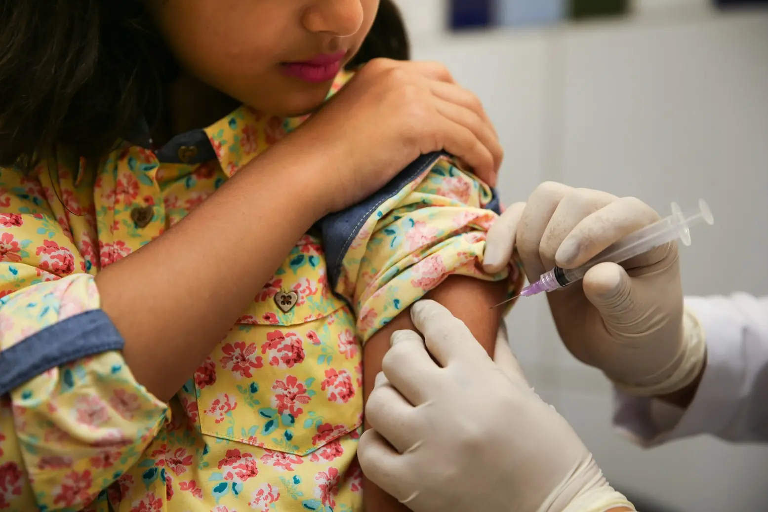 Uma criança recebe uma injeção no braço de um profissional de saúde usando luvas. A criança veste uma camisa floral amarela.