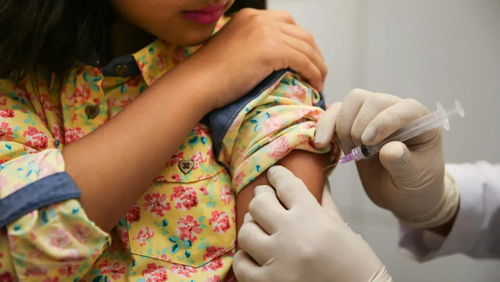 Uma criança recebe uma injeção no braço de um profissional de saúde usando luvas. A criança veste uma camisa floral amarela.