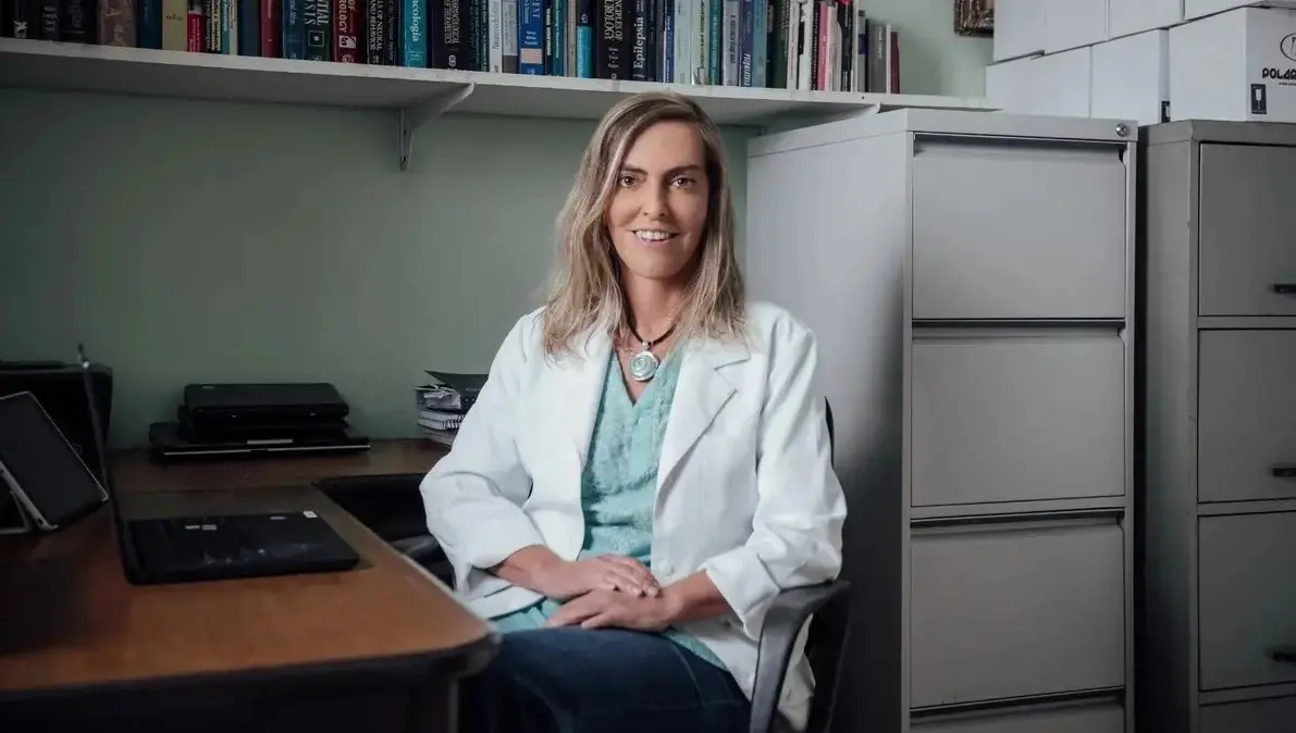 Tatiana é uma mulher de meia idade, branca, cabelo loiro tamanho médio, liso. Na foto, ela está de jaleco branco, blusa verde e calça jeans, com as mãos sobre as pernas.