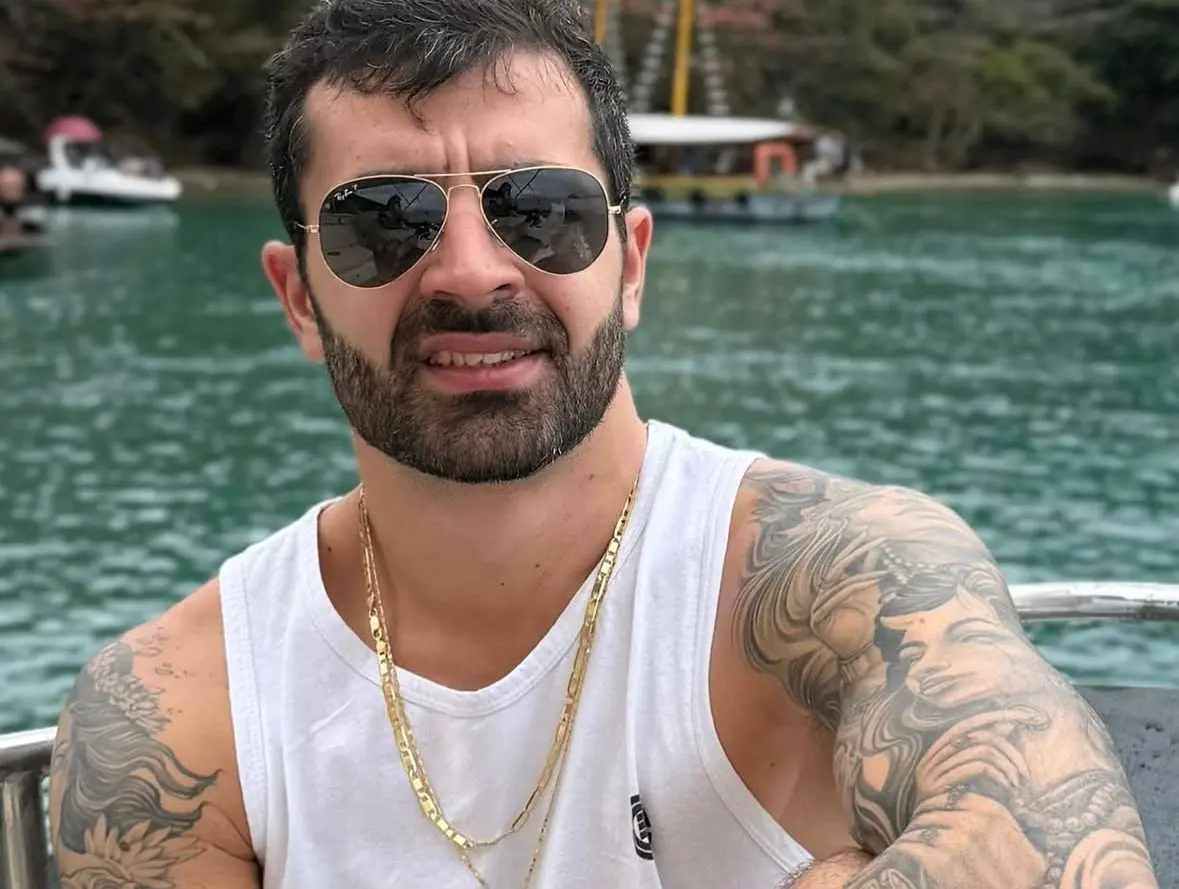 Bruno é um homem branco de barba preta bem feita e cabelo curto da mesma cor. Na foto, ele está de regata branca e usa um colar dourado. Tem tatuagens pelo braço e está de óculos escuros.