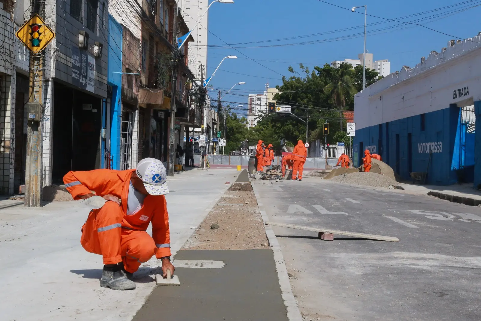 Obra de pavimentação de ruas por trabalhadores em uniforme laranja na cidade, com construções e edifícios ao fundo, serviço de revitalização urbana em andamento.