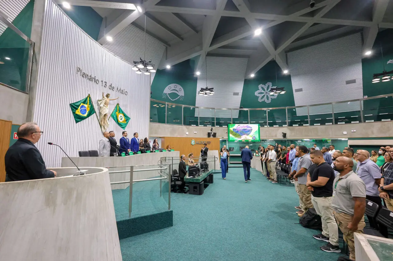 Interior de uma assembleia, com duas bandeiras do Brail ao centro do local e múltiplas pessoas em pé durante sessão.