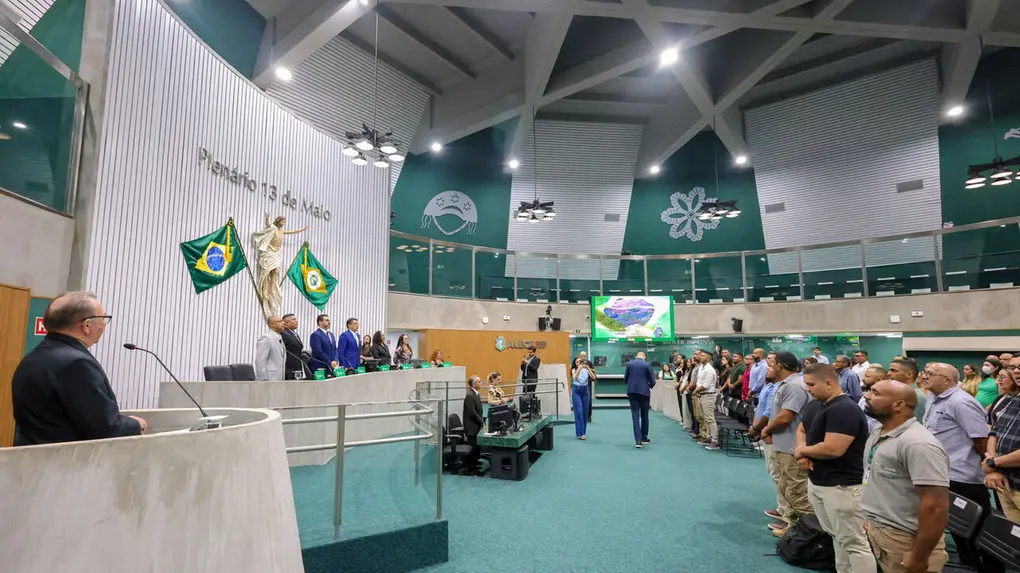 Interior de uma assembleia, com duas bandeiras do Brail ao centro do local e múltiplas pessoas em pé durante sessão.