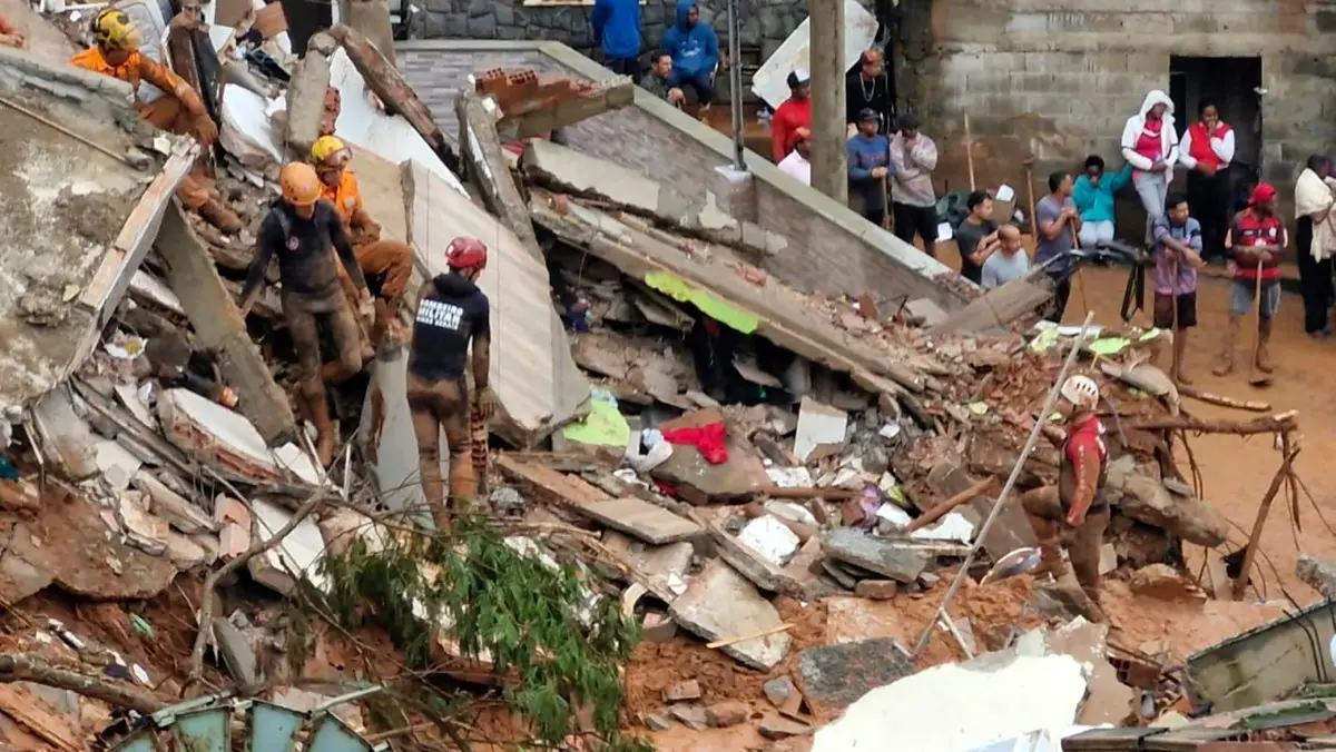 Equipes de resgate trabalham sobre os escombros de estruturas destruídas após um deslizamento de terra. Diversos profissionais com capacetes e roupas de proteção procuram por vítimas entre pedaços de concreto, madeira e lama. Ao fundo, moradores observam a operação próximos a casas danificadas.