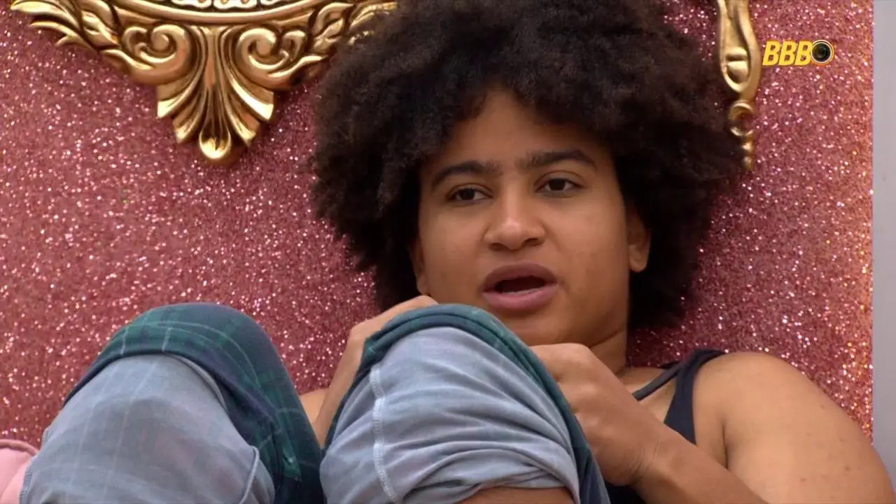 Close-up de uma pessoa com cabelo crespo escuro, sentada em um ambiente com parede brilhante rosa e detalhes dourados, com o logotipo do BBB no canto superior direito. Parece estar ouvindo ou em meio a uma fala.