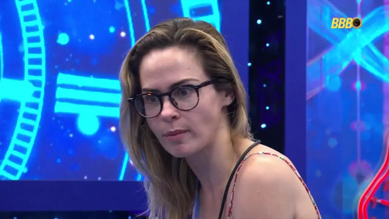 Ana Paula, mulher de cabelo loiro-escuro e óculos, com expressão séria, em close-up, contra um fundo azul vibrante com padrões luminosos abstratos e o logo do BBB.