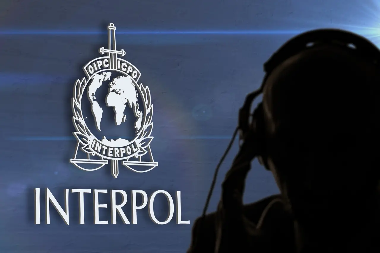 Logotipo da Interpol com o emblema do globo e a balança sobre fundo azul, ao lado da palavra “INTERPOL” em destaque. À direita, há a silhueta de uma pessoa usando fones de ouvido, parcialmente desfocada.
