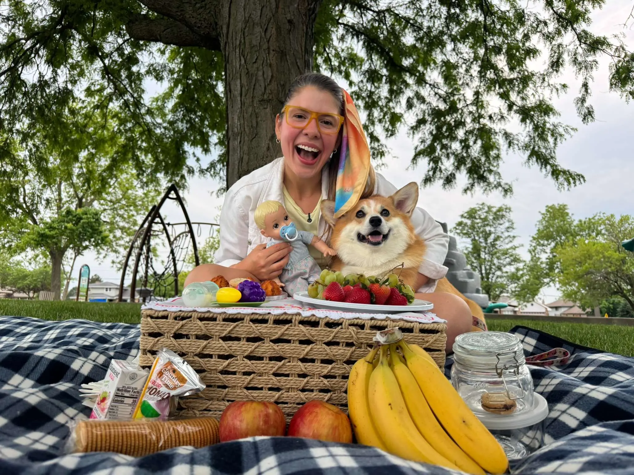 Uma jovem sorridente de óculos, segurando uma boneca, sentada em um piquenique com um cachorro Corgi feliz, frutas e lanches sobre um cobertor xadrez azul..